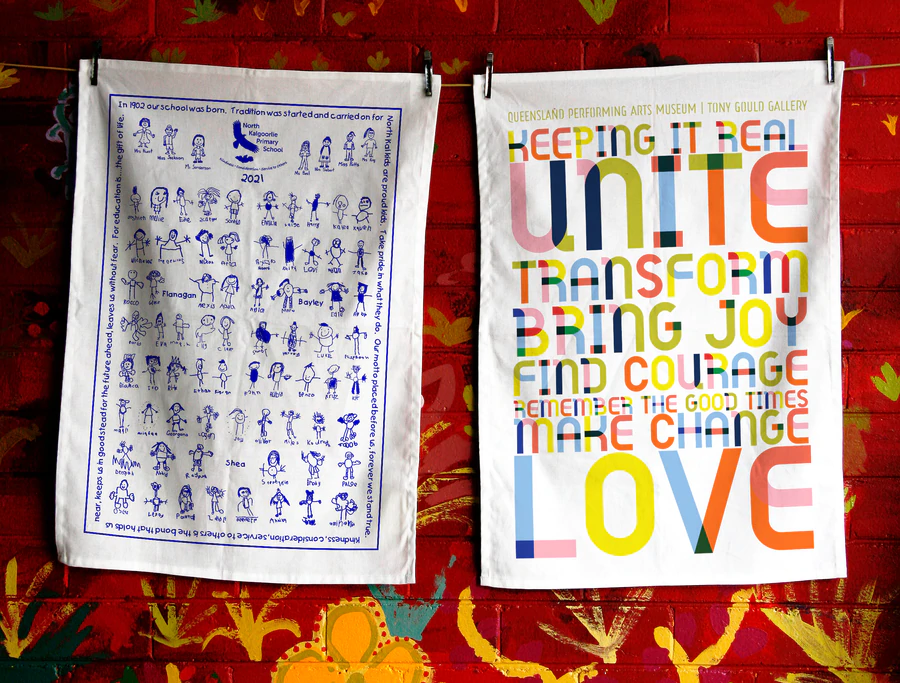 Two custom printed tea towels from Expressions Fundraising. The left features blue child-like drawings and a school song border by North Kalgoorlie Primary School for a 2021 school tea towel fundraising project. The right shows a colourful typographic design with uplifting words by Queensland Performing Arts Museum, used as ethical promotional merchandise.