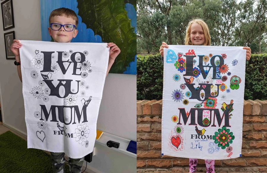 Children holding customised “I Love You Mum” tea towels, one uncoloured and one fully coloured.
