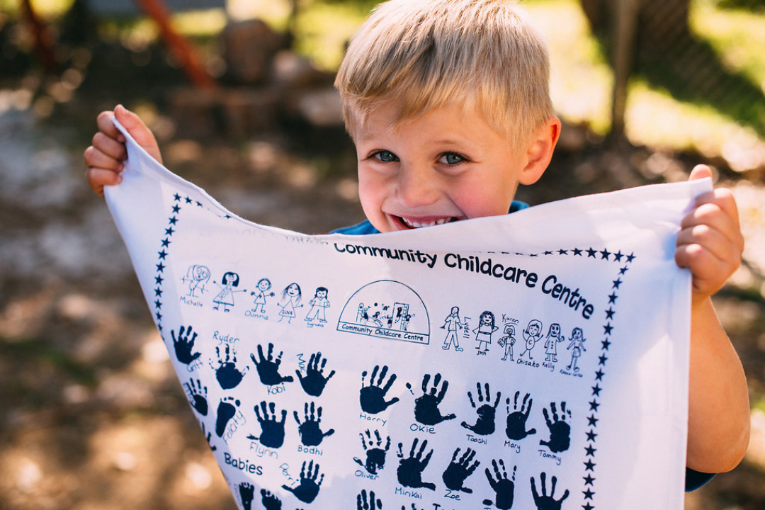 A smiling young boy holding up a printed tea towel featuring hand-drawn portraits, handprints, and names of children from a Community Childcare Centre.