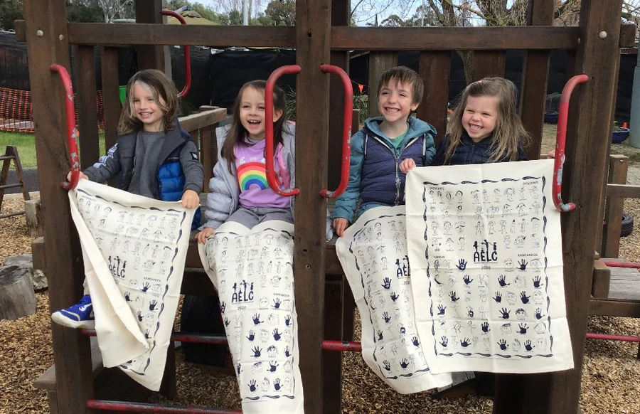 Four smiling children from Armadale Early Learning Centre sitting on a playground structure, proudly holding up custom printed organic cotton tea towels featuring their handprints and self-portrait drawings as part of their school tea towel fundraising project.