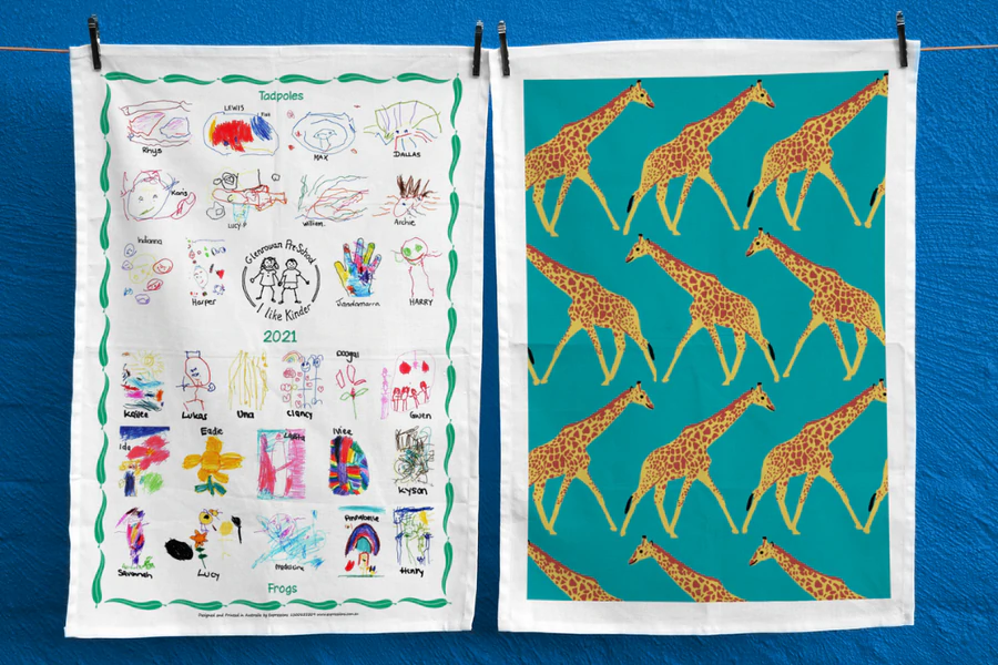 Two tea towels on display—one featuring children’s drawings from Glenrowan Preschool, the other with a repeating giraffe illustration.