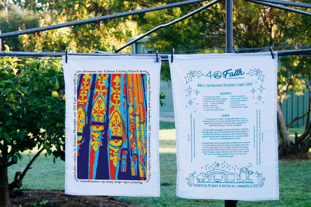 Two custom printed tea towels hanging outdoors on a clothesline. The left tea towel features a vibrant stained glass organ pipe design from Galston Uniting Church. The right tea towel showcases a recipe for Oma’s Apfelkuchen (Grandma’s Apple Cake) with decorative text and illustrations, commemorating 40 years of Faith Lutheran College.