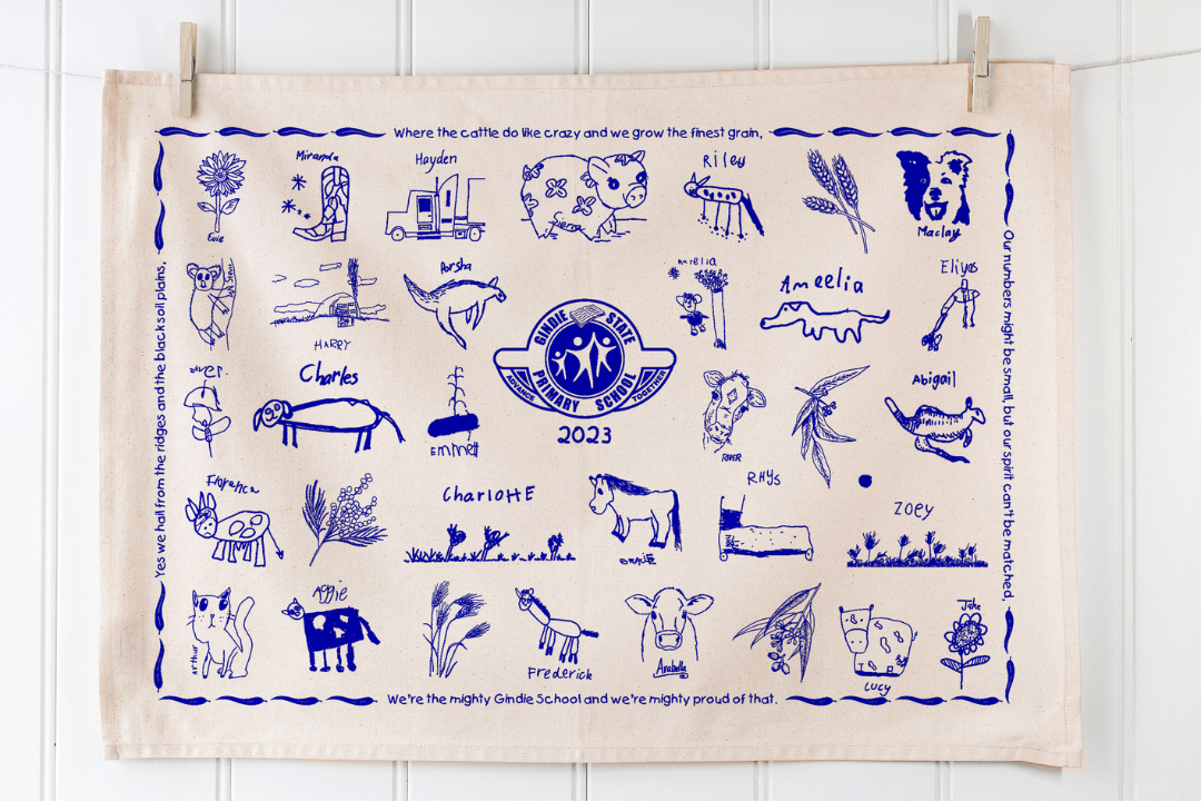Hand-drawn children's illustrations printed in blue on a cream tea towel from Gindie State Primary School 2023, featuring names and artwork arranged around a central badge. Displayed vertically against a white wall, this creative and sentimental design showcases the charm of personalised school fundraising with Expressions.