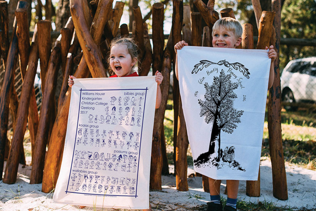 Two smiling children holding custom tea towels featuring school artwork – Expressions Fundraising keepsakes made in Australia