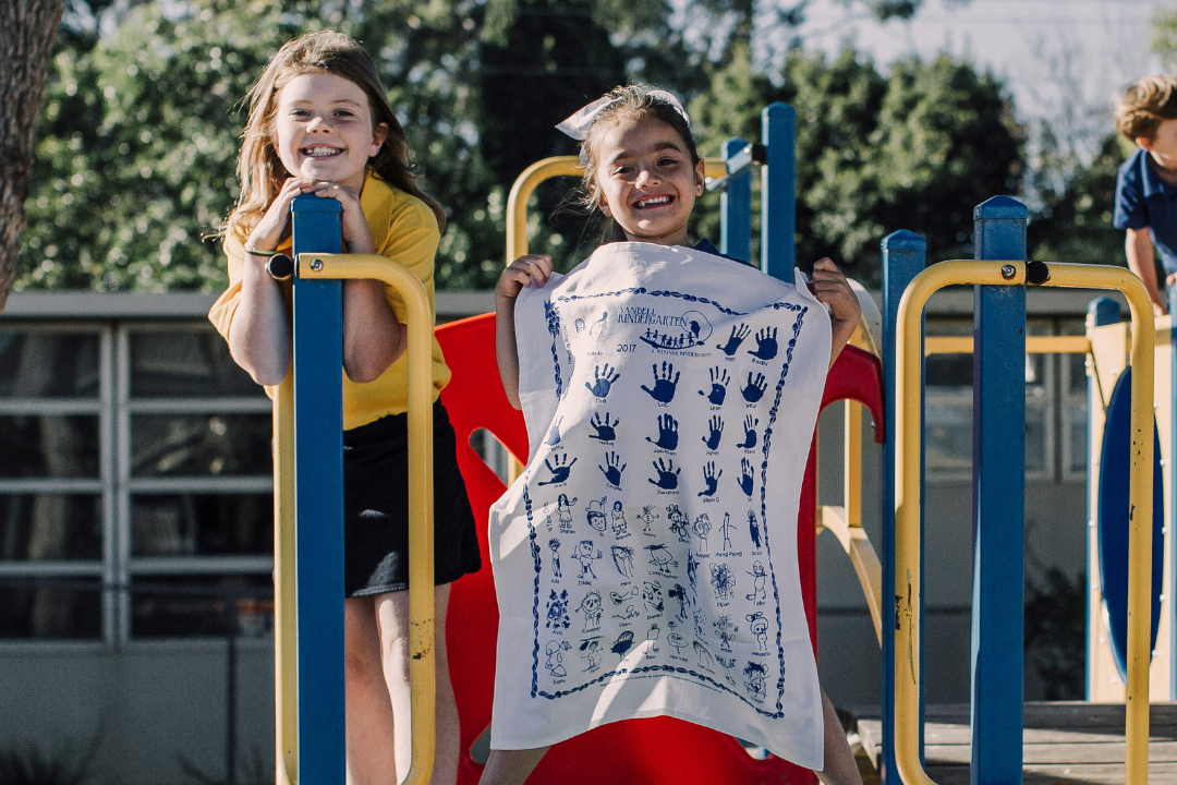 Two smiling schoolgirls on a playground slide, one proudly holding a personalised tea towel with handprints and drawings—Expressions Fundraising 2025.