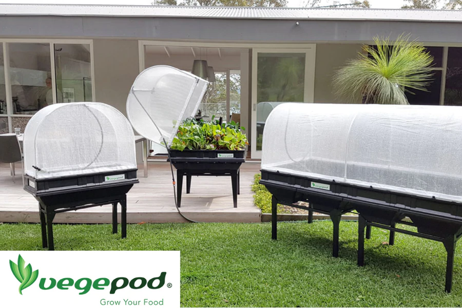 Three Vegepod raised garden beds with protective mesh covers set up in a lush backyard, one filled with growing vegetables. Logo of Vegepod with the tagline ‘Grow Your Food’ displayed.