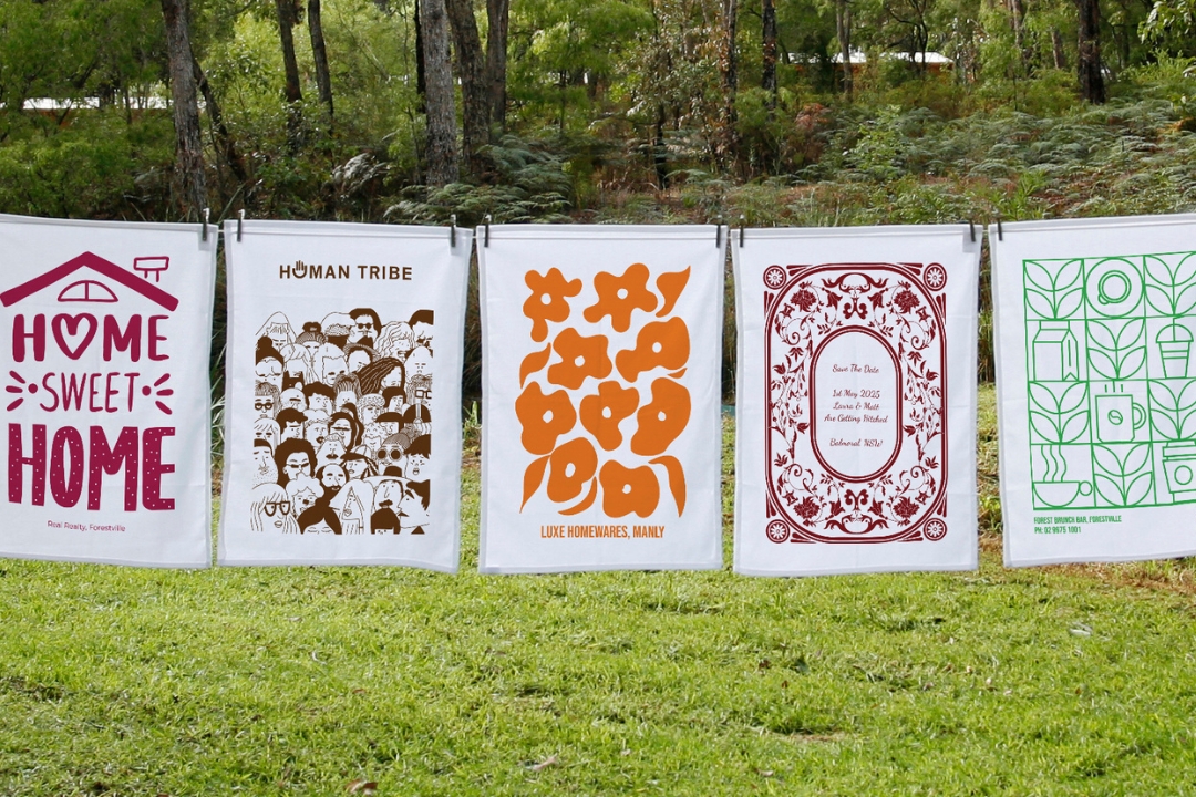 Five custom-printed tea towels from Expressions Australia displayed on a clothesline outdoors, showcasing creative community artwork and personalised designs.