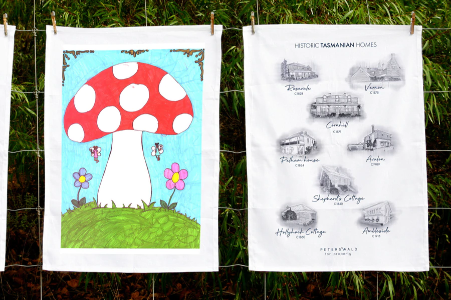 Two custom printed tea towels hanging on a wire fence. The left towel features a colourful hand-drawn red and white mushroom with flowers and butterflies, designed by a child. The right towel showcases monochrome illustrations of historic Tasmanian homes with detailed labels, including names like Roseneath, Vernon, and Shepherd's Cottage.