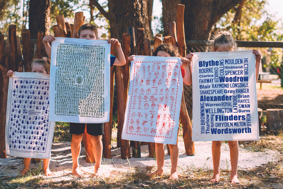 Four young children proudly holding up custom printed tea towels outdoors. Each tea towel features unique school or childcare-themed artwork including handprints, student self-portraits, and bold text designs, celebrating creative and sustainable fundraising through Expressions Fundraising.
