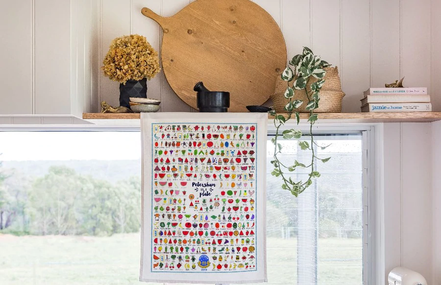 Colourful tea towel titled “Petersham on a Plate” hanging in a sunlit kitchen with wooden and botanical decor.