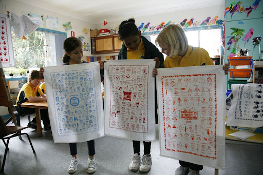 Three school students proudly holding up their Expressions Tea Towel fundraiser designs in a colourful classroom, each featuring student artwork in blue, red, and orange prints.