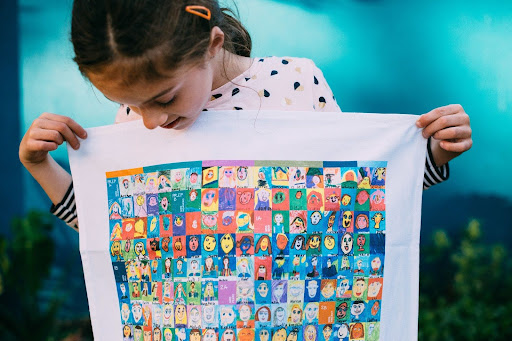 Young girl proudly holding a colourful school tea towel featuring a grid of hand-drawn self portraits by students from multiple grades, printed on white cotton fabric.
