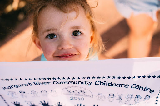 Young child smiling while holding a custom printed tea towel from Margaret River Community Childcare Centre featuring student drawings