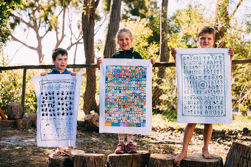 Three children proudly holding up custom Expressions Tea Towels featuring colourful and hand-drawn designs from their school or childcare centre, outdoors on a sunny day.