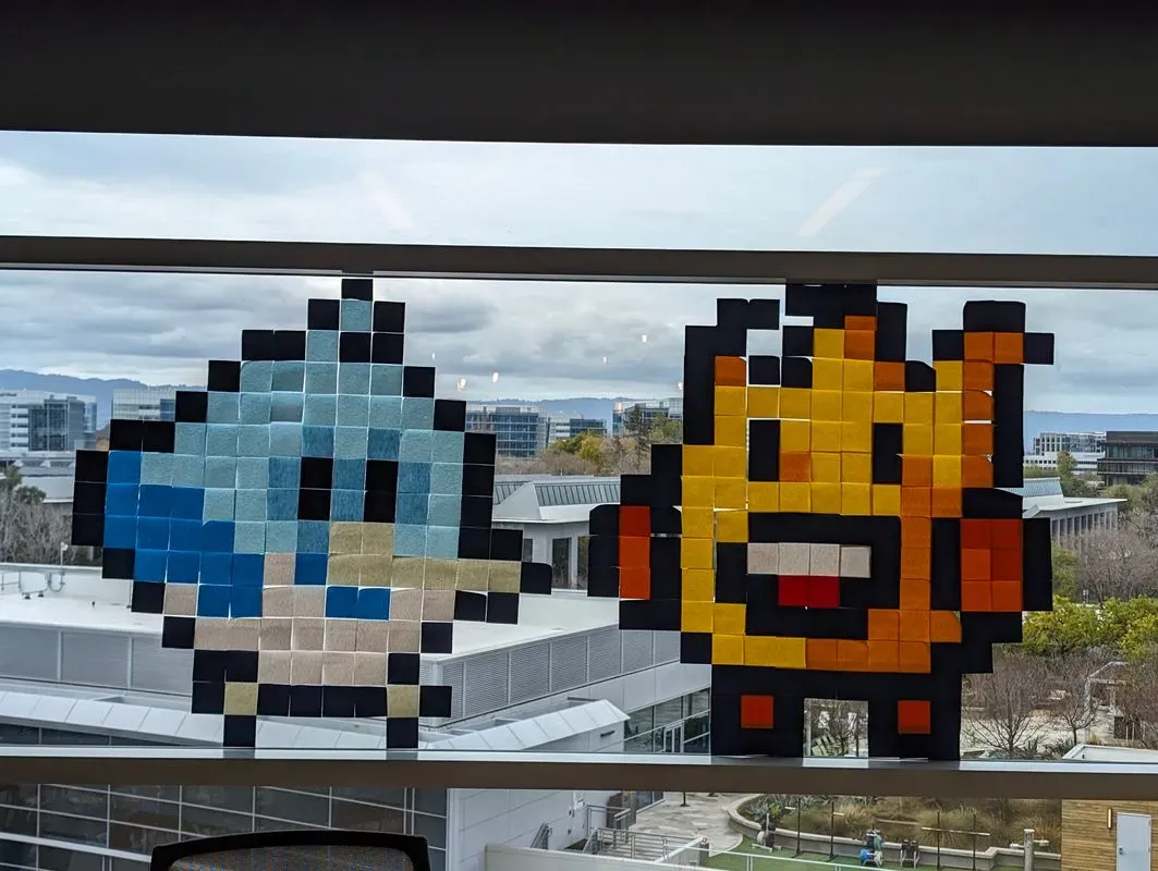 Two pixel art characters made of sticky notes are attached to a window. The character on the left is blue and the one on the right is yellow.