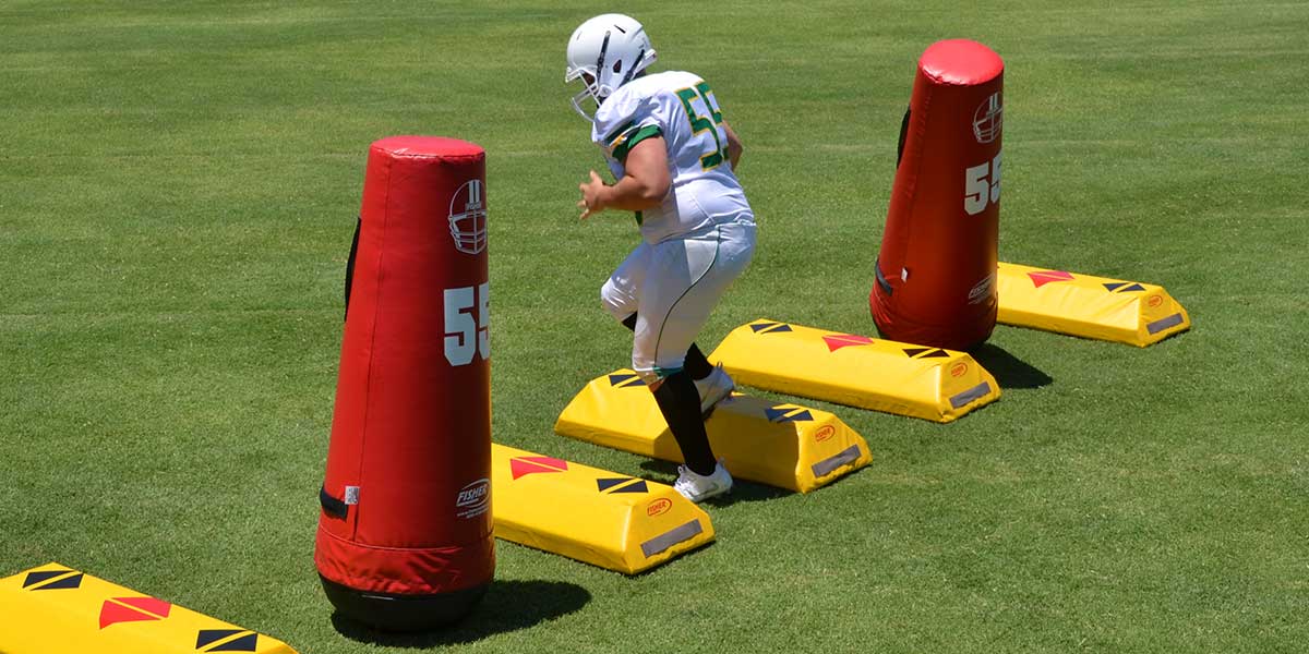 Two red Fisher Athletic Varsity Pop-Up Dummies standing on the ground in a close-up shot. A player is practicing lateral movement, maneuvering around the dummies during a training drill.