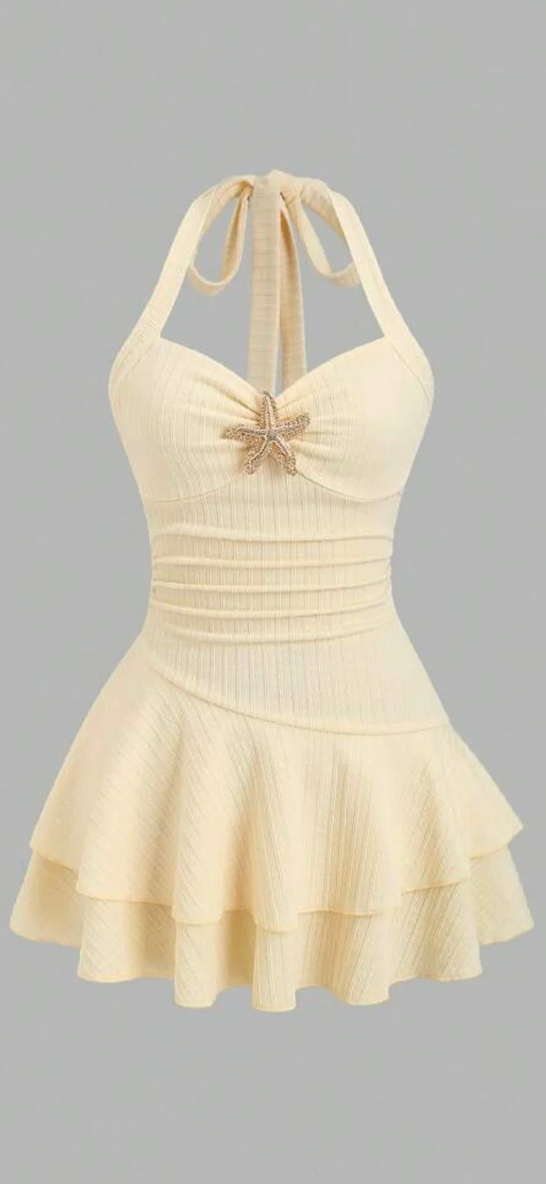 A vintage-style cream-colored swimsuit with a peplum design. The top portion features a halter neck design with a tie at the back of the neck. The bodice has a sweetheart neckline with a decorative starfish brooch in metallic gold positioned at the center. The fabric has a textured, ribbed pattern throughout. The peplum waist creates a fitted silhouette with a layered ruffle skirt that extends to mid-thigh length. The skirt portion has two tiers of ruffles that create a flared, feminine silhouette. The swimsuit is photographed against a plain light gray background. The garment is displayed as if worn, showing natural draping and the full design of the piece.