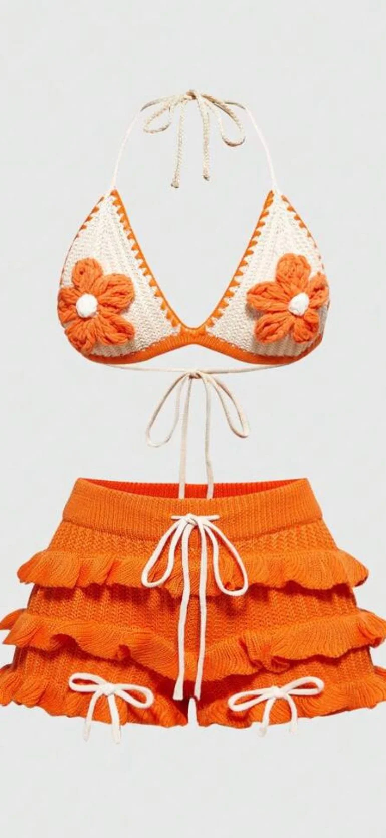 A vintage-style two-piece swimsuit set photographed against a plain white background. The top piece is a triangle-style bikini with a white base color and orange trim. Two large crocheted orange daisy flowers with white centers are attached to the top. The bikini straps are thin and white, tied in the back. The bottom piece is a high-waisted ruffled skirt in solid orange with three tiers of ruffles. White drawstrings are attached at each side of the ruffled skirt for adjustment. The ruffles have a textured, crocheted appearance. The swimsuit is photographed in a flat lay style, with the top piece positioned above the bottom piece. The image has a clean, minimalist aesthetic with sharp contrast between the white background and the orange and cream-colored swimwear.