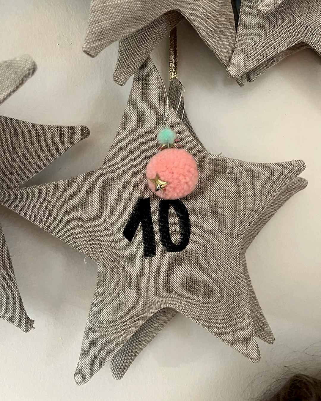 Detalle de una estrella de lino gris con el número 10 pintado en negro y un pompón rosado con una estrella dorada, colgada de una cinta
