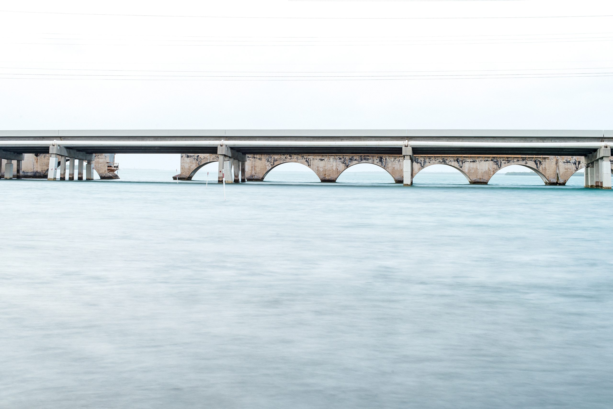 A modern highway overpass spans calm turquoise water, its concrete deck supported by five large cylindrical pillars rising from the seafloor. Weathered stone structures beneath the upper roadway suggest older infrastructure, possibly fortifications or earlier construction.