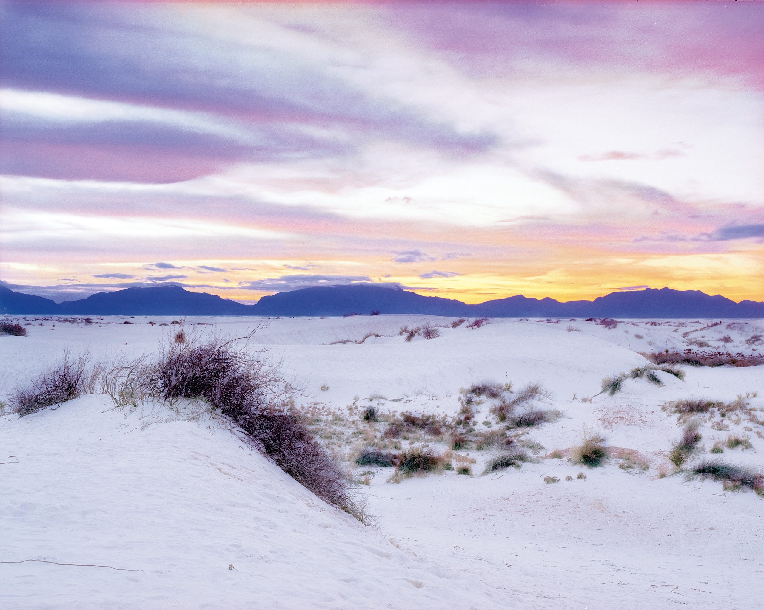 White sand dunes with sparse desert vegetation stretch toward a mountain range silhouetted against a twilight sky of pink, purple, and gold. The sun sets behind the distant peaks while clouds catch the last light overhead.