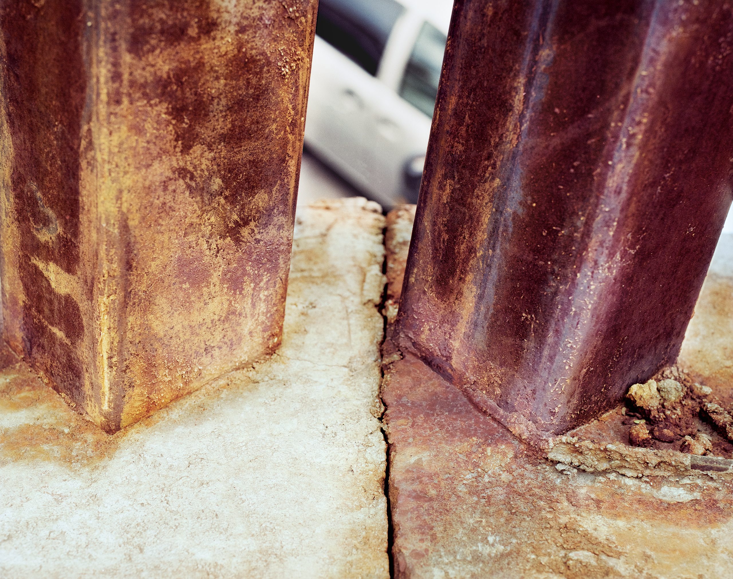 Two heavily rusted metal posts stand in a concrete foundation, their surfaces oxidized to deep reddish-brown. Loose rust debris and concrete dust accumulate at their bases.