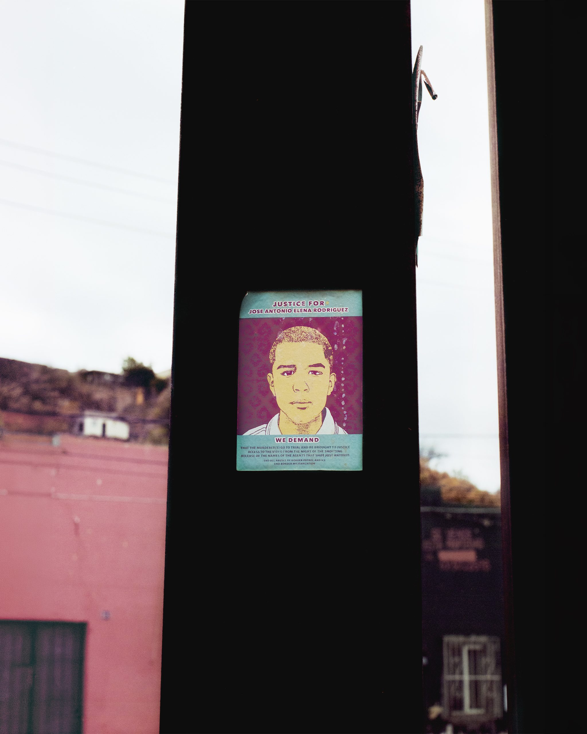 A printed poster affixed to a dark metal beam shows a portrait illustration with the text "Justice for Jose Antonio Elena Rodriguez" and "We Demand" above and below. Behind the beam, a pink concrete building and desert hills are visible in soft afternoon light.