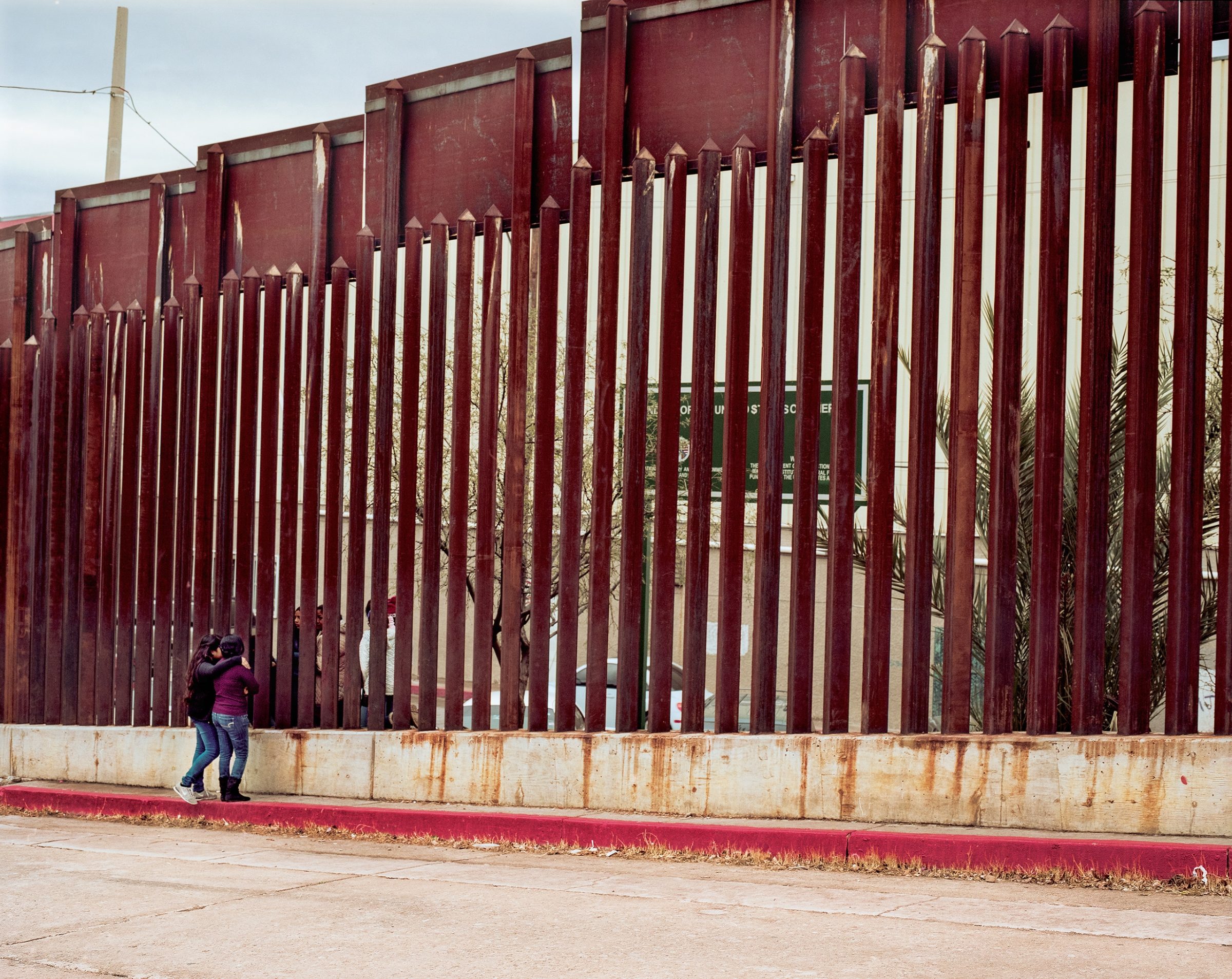 A person in a maroon jacket stands before a massive corrugated metal border fence, its tall vertical slats painted deep red, with rust staining the concrete foundation below. The fence recedes into the distance under overcast sky, with industrial structures visible beyond.