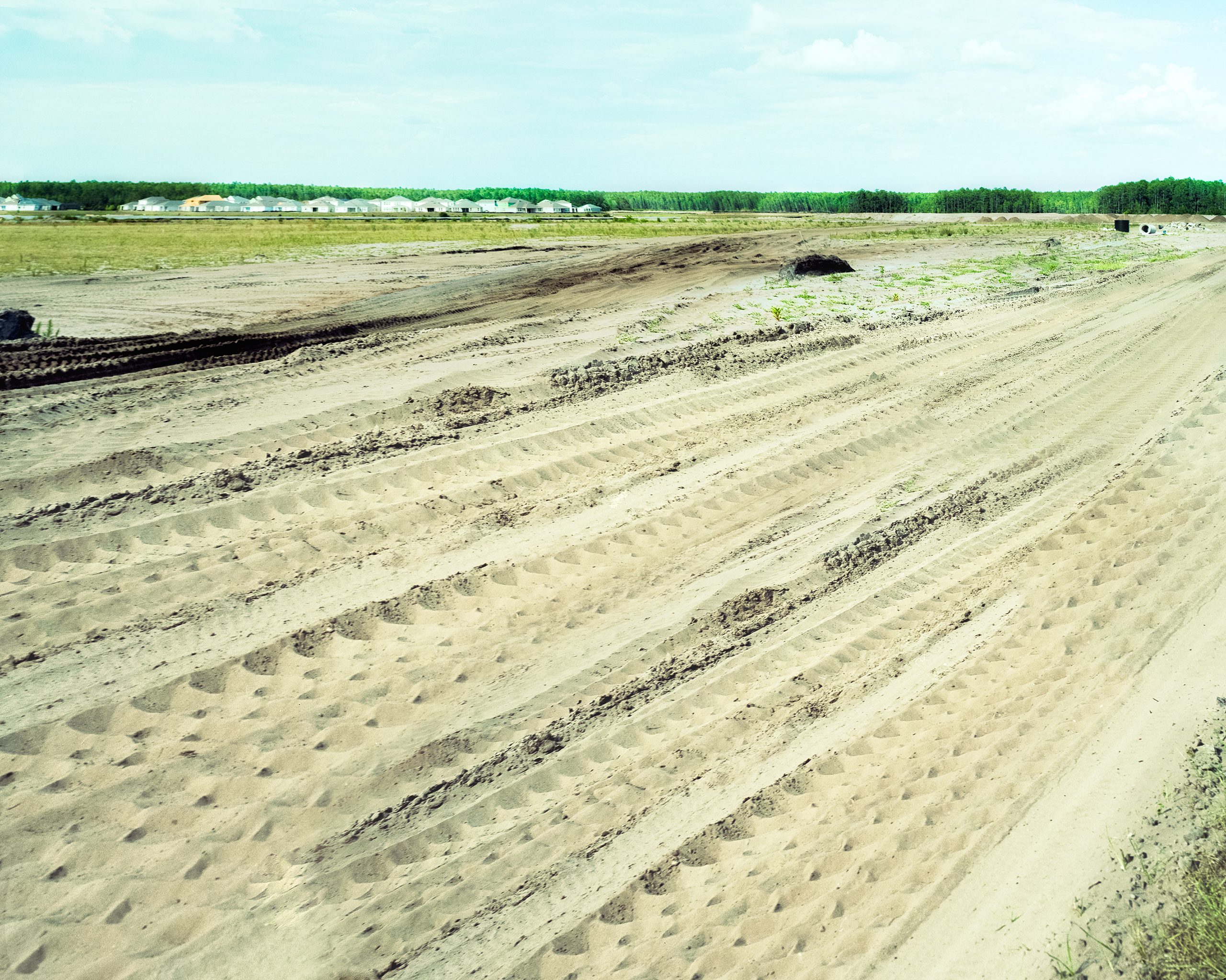 Furrowed sandy field in the foreground with planted rows receding toward a tree line, farm buildings and structures visible along the distant horizon under a pale blue sky.