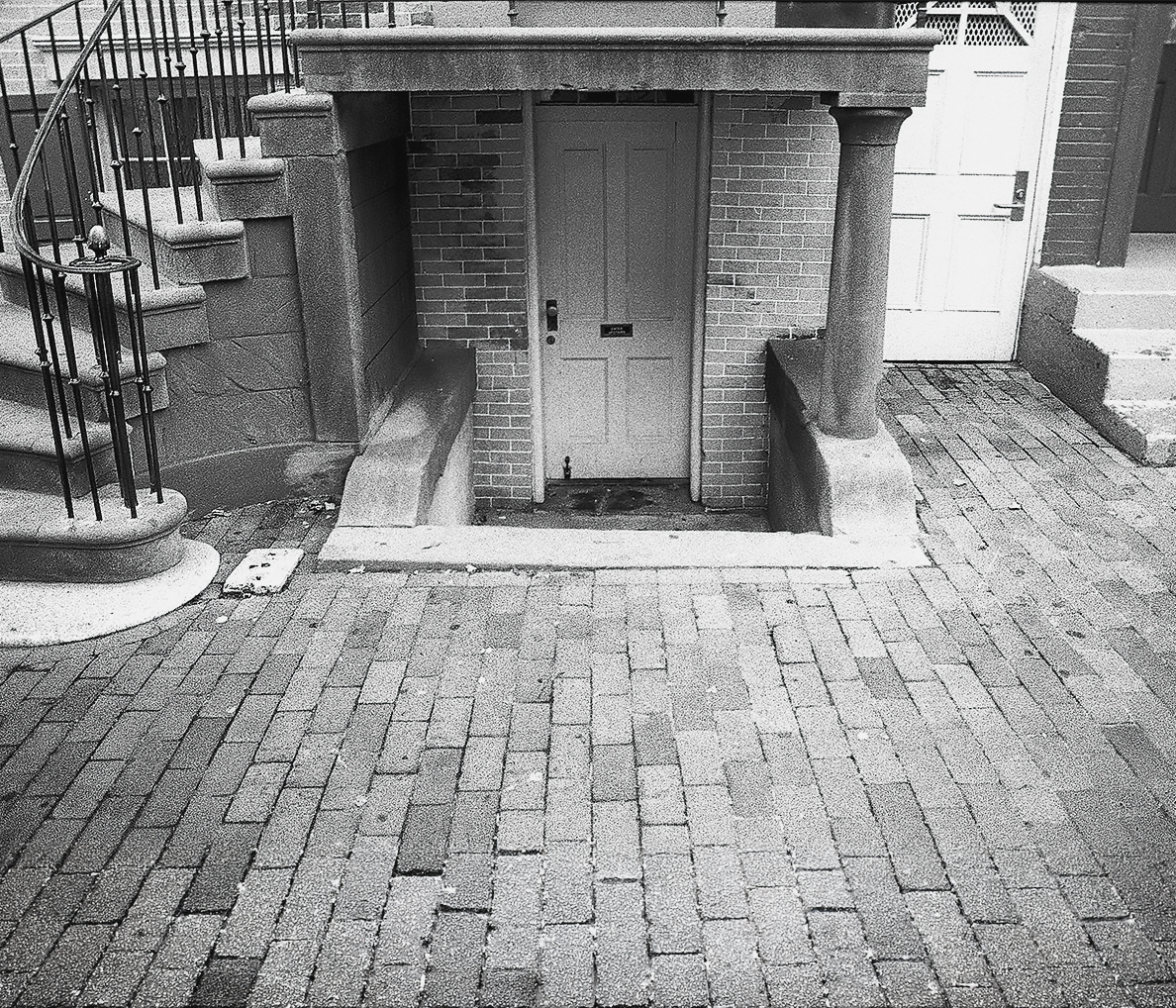 A sunken basement entrance with brick walls and a metal door recessed below street level, flanked by concrete pillars. A spiral staircase with metal railings descends on the left, and herringbone-patterned pavement surrounds the entryway.