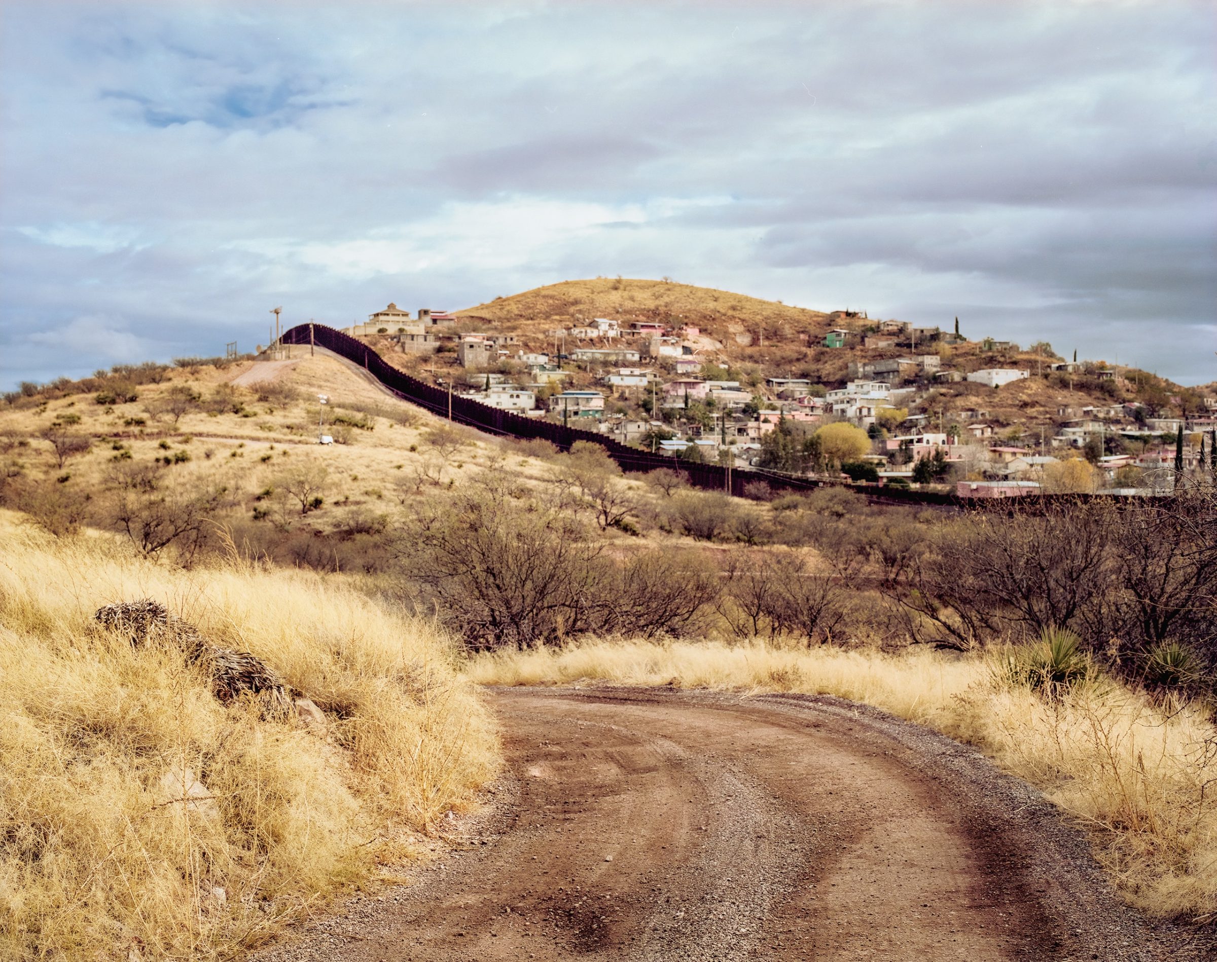 A dirt road curves through dry grassland toward a hillside town where houses cluster around the slope, with a border wall dividing the desert landscape in the foreground under an overcast sky.