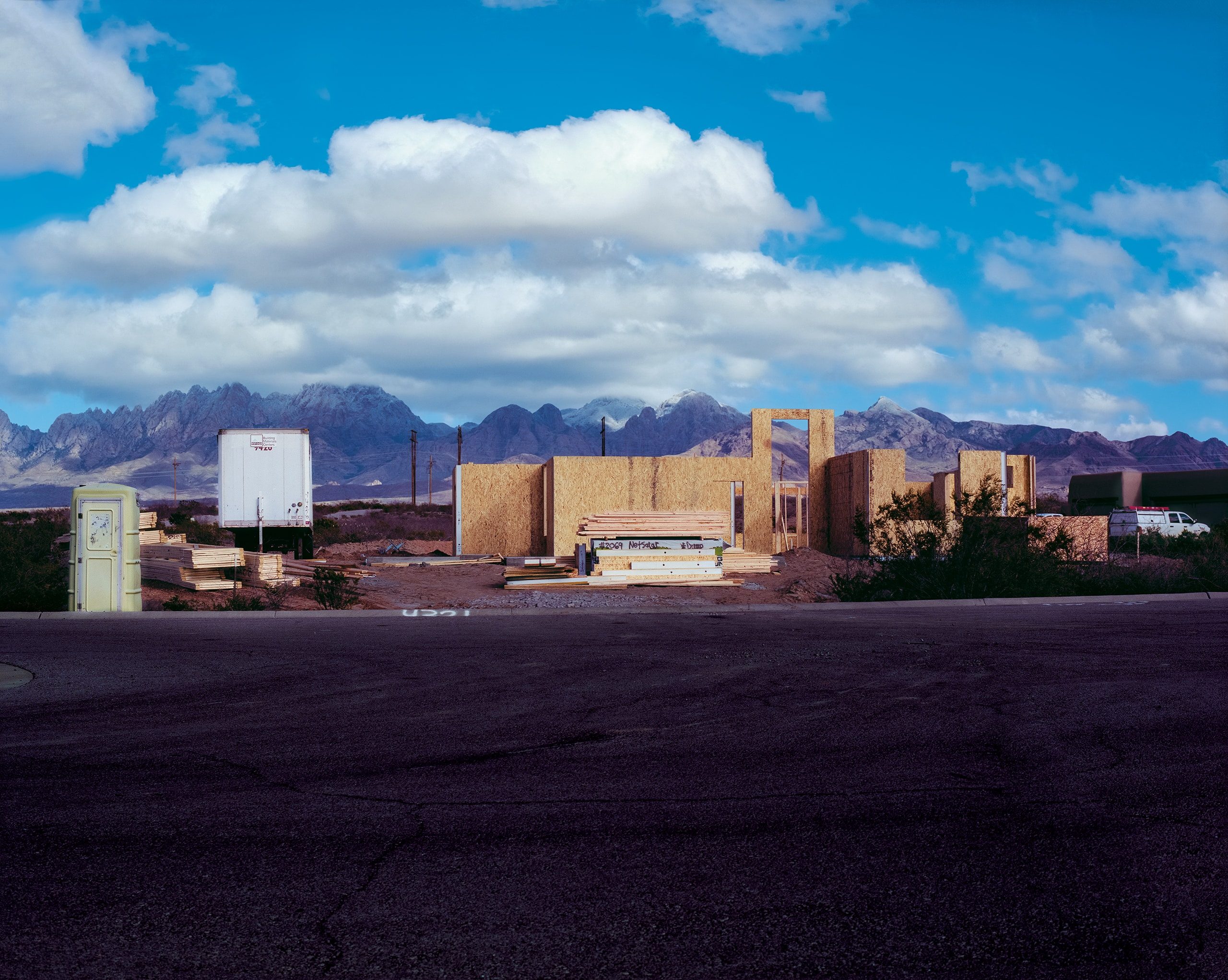 A construction site in the desert with plywood walls and framing materials stacked beside a portable toilet and white storage unit, mountains rising sharply in the distance under a bright sky.