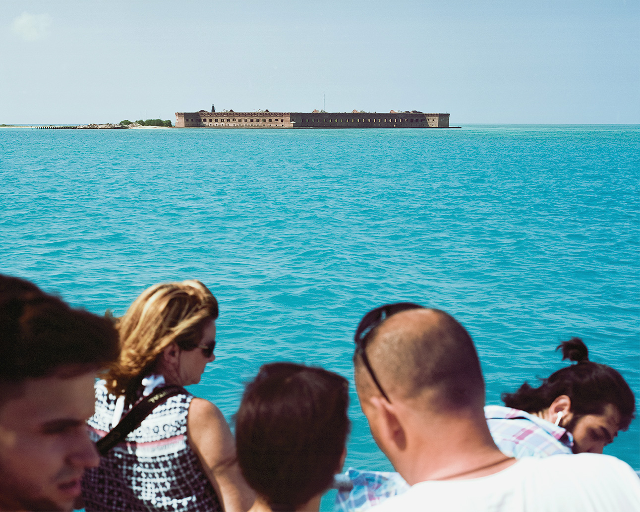 Tourists on a boat observe Fort Jefferson, a massive red-brick military fortress on a small island surrounded by turquoise water under clear sky.
