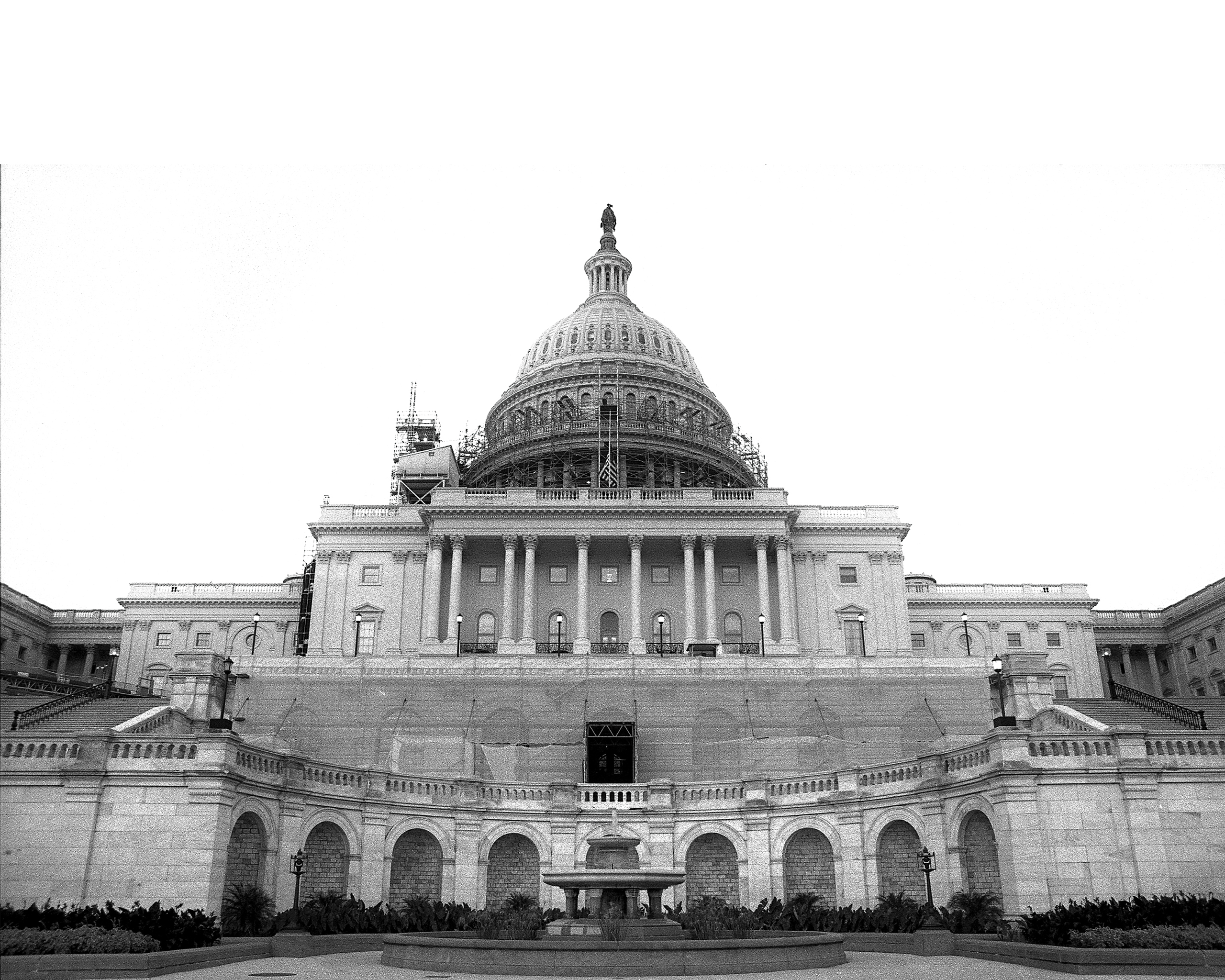 The U.S. Capitol building's east facade in black and white, viewed from the plaza below. Scaffolding covers the dome, and a temporary structure obscures the lower entrance during renovation work.