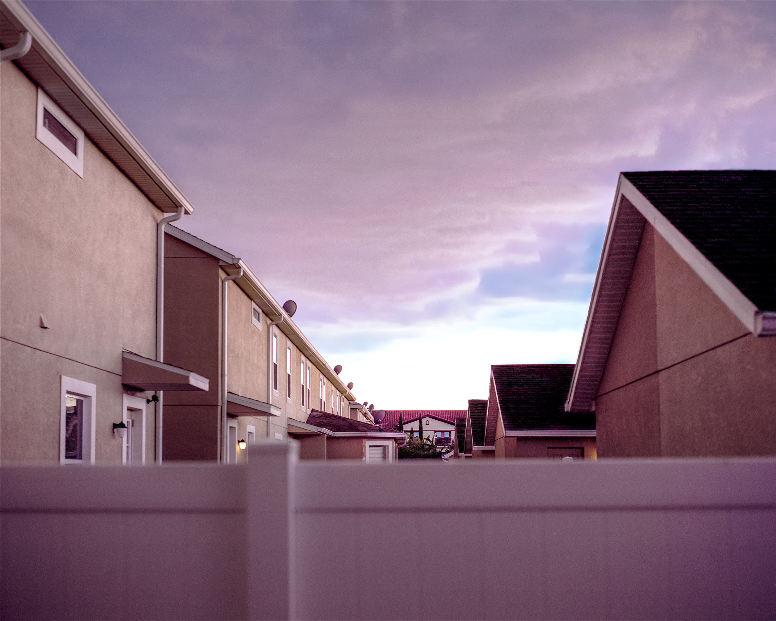 A row of connected residential buildings with brick and stucco facades stretches across a paved courtyard at dusk, their rooflines silhouetted against a purple and pink sky. The structures appear to be modest housing units or townhouses, photographed from ground level looking slightly upward.