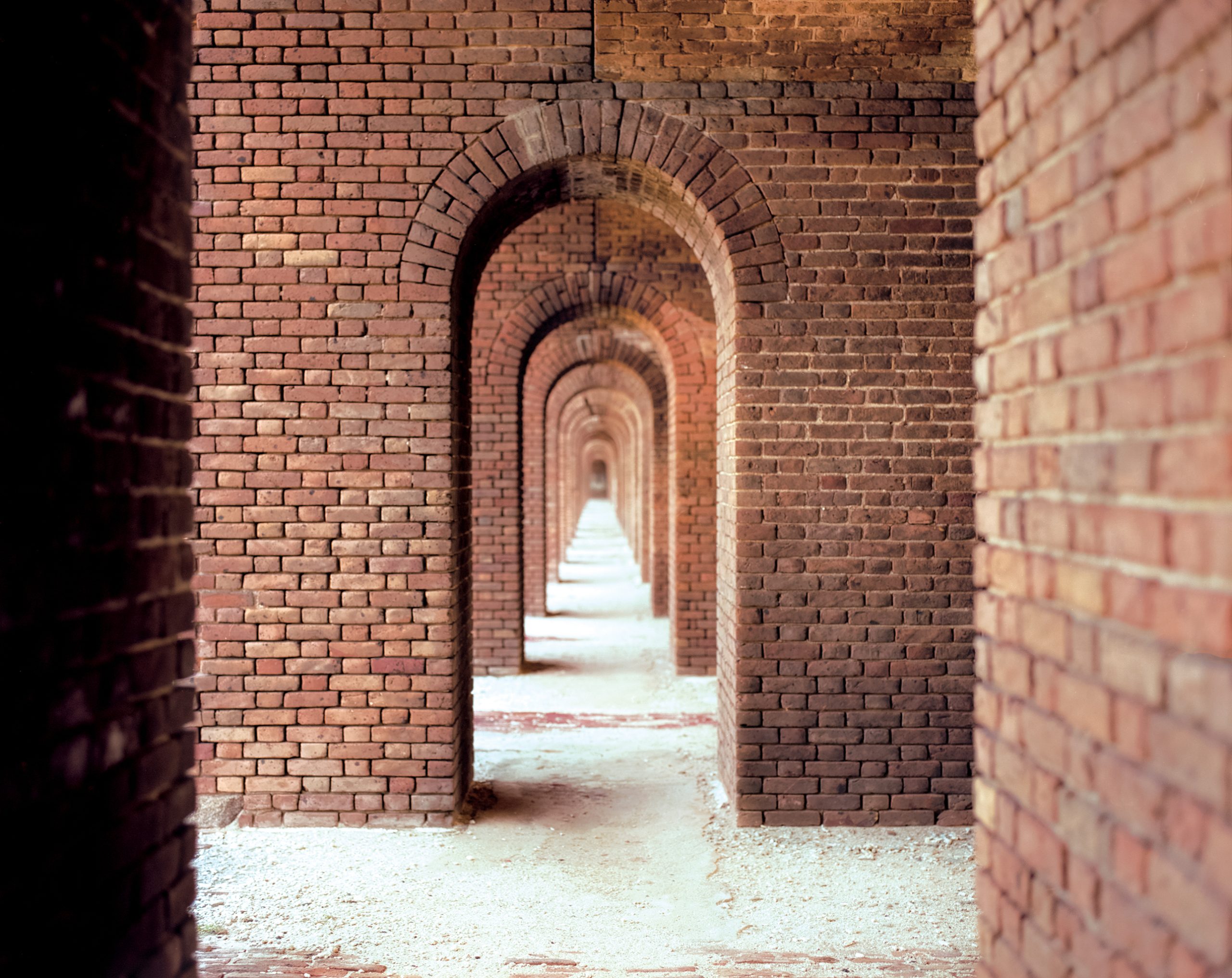 A succession of red-brick arches recedes into the distance through an empty corridor, their curves creating a tunnel effect. Sunlight enters from the far end, illuminating the sandy floor and the precisely laid brickwork of the vault.