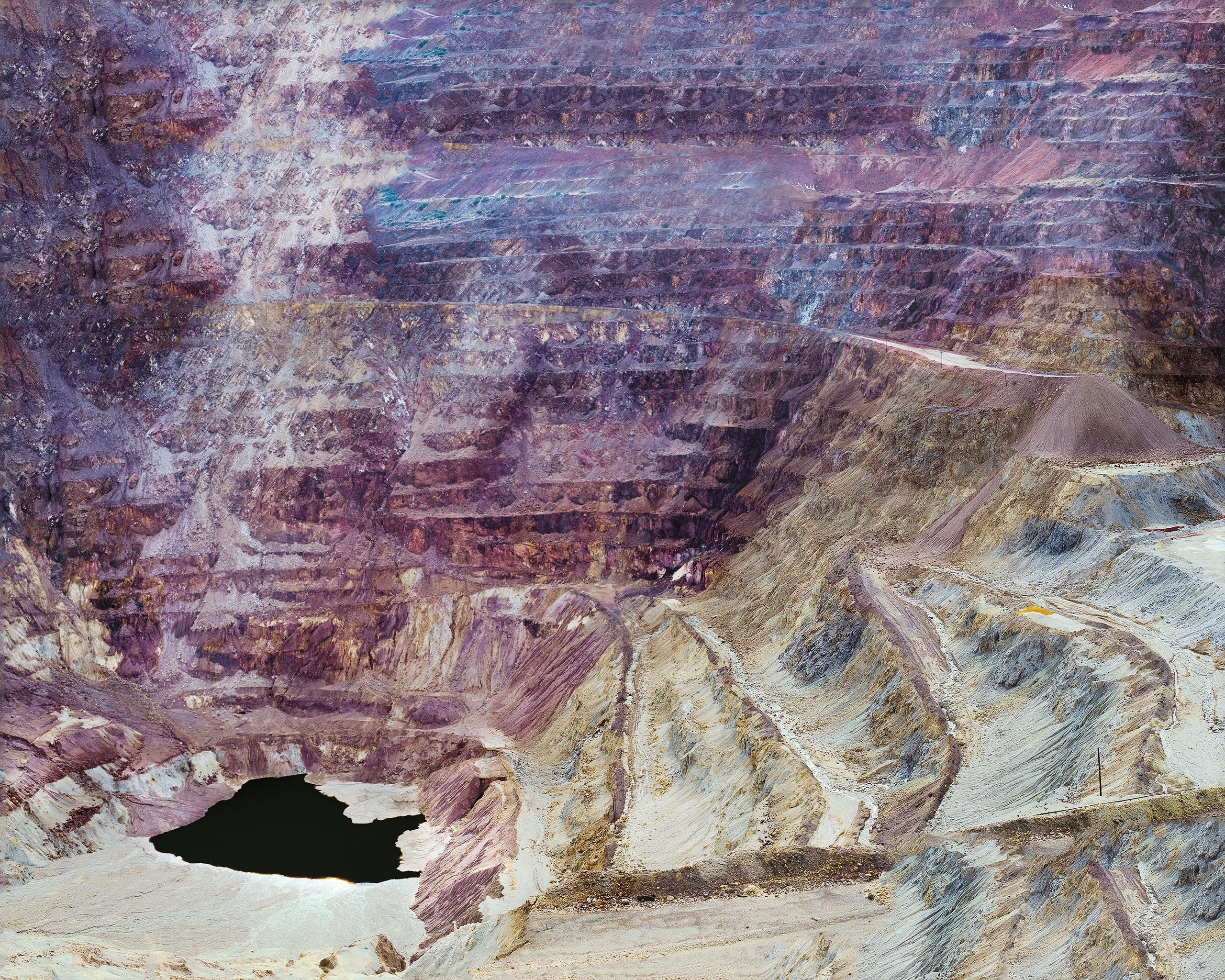 A deep open-pit mine with layered purple and rust-colored rock faces descends in geometric terraces. A dark pool of water sits at the bottom left, while a pale tailings pile and access road occupy the lower right slope.