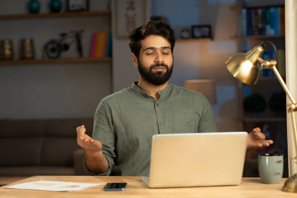 Young Indian man practicing breathing exercises with eyes closed while sitting at a desk with a laptop, coffee mug, and documents, creating a calm and focused work-from-home environment.