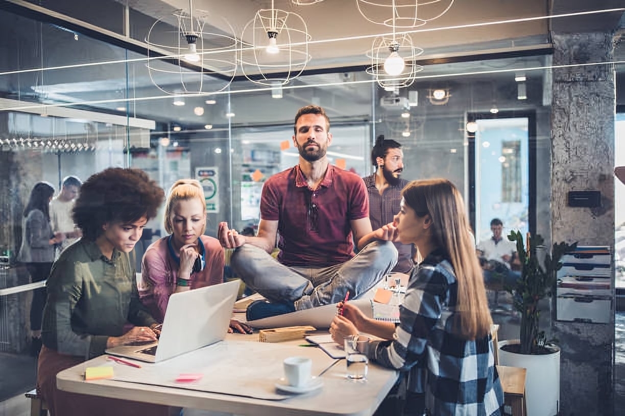 Man practicing breathing exercises on a table in a modern office while coworkers work around him, promoting stress relief at work.