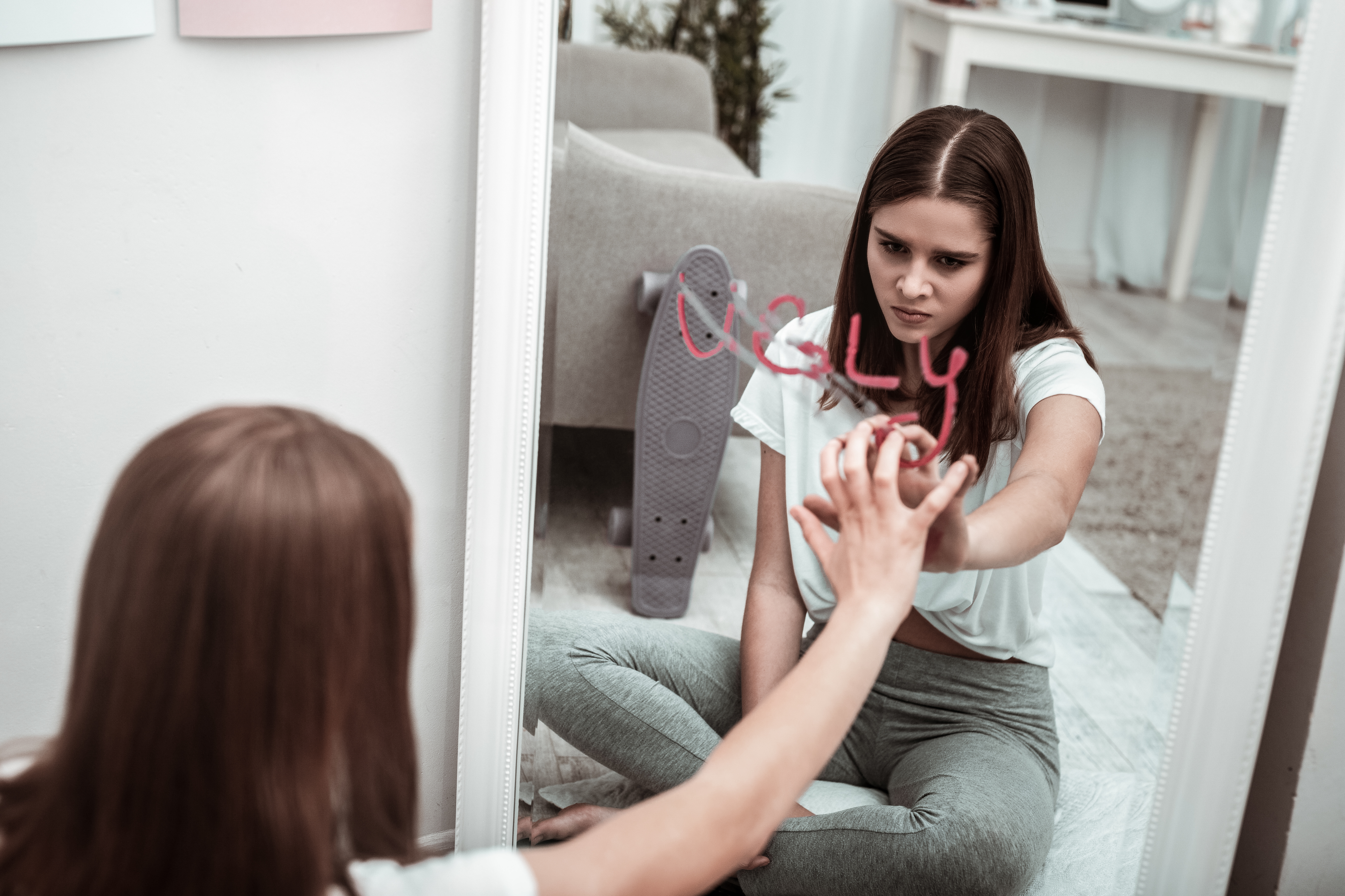 Teen girl struggling with low self-esteem looking into mirror with "ugly" written on it – concept of self-hatred 