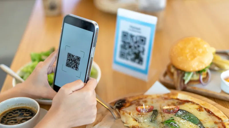 Código QR impreso sobre una mesa o mostrador, alguien escaneándolo con el celular