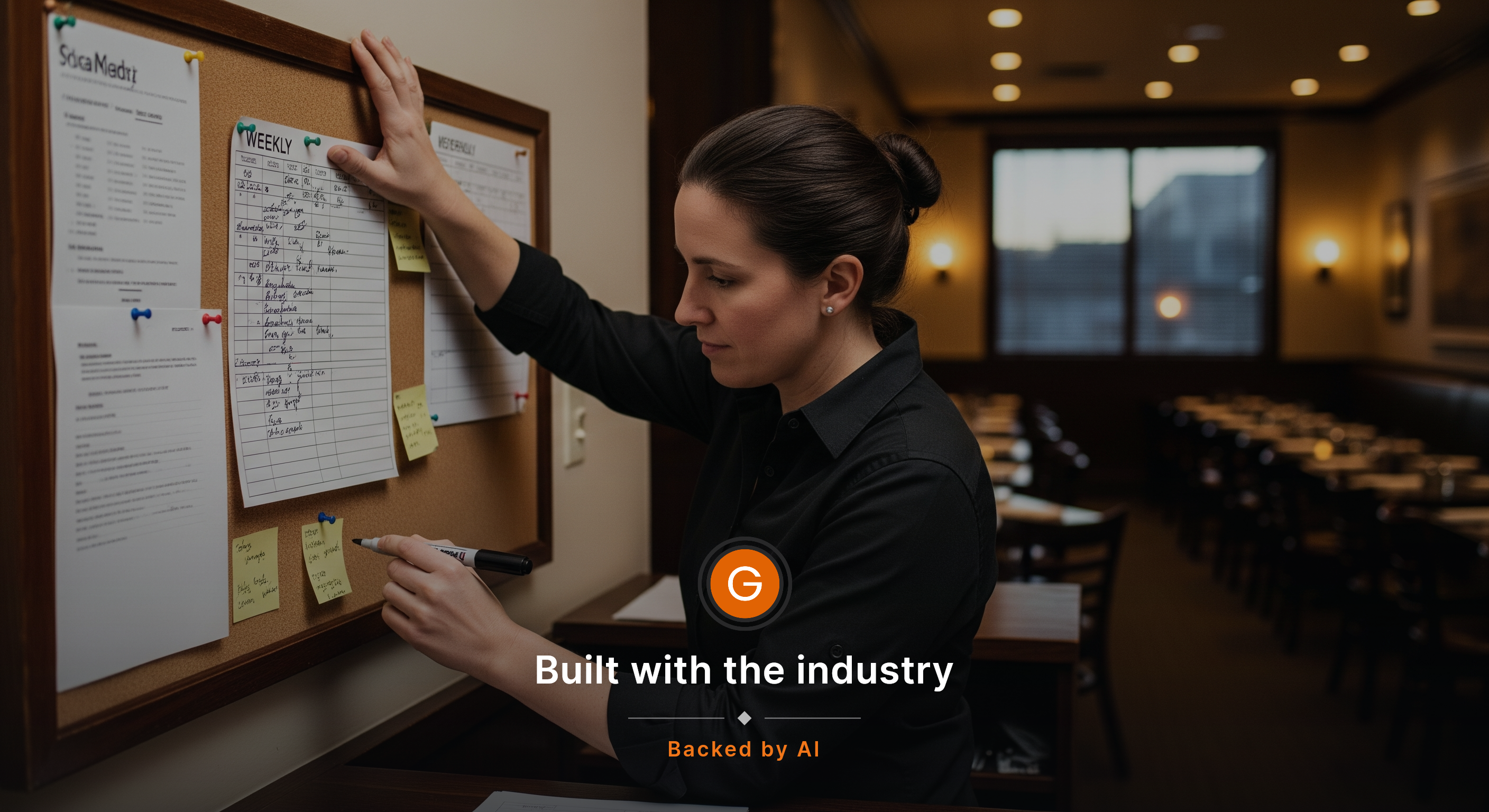 An environmental portrait of a restaurant manager alone at the host stand before service, reviewing a handwritten weekly schedule pinned to a corkboard, one hand on the board and the other holding a marker, dining room softly lit behind them. The worker is focused and not looking at the camera, with