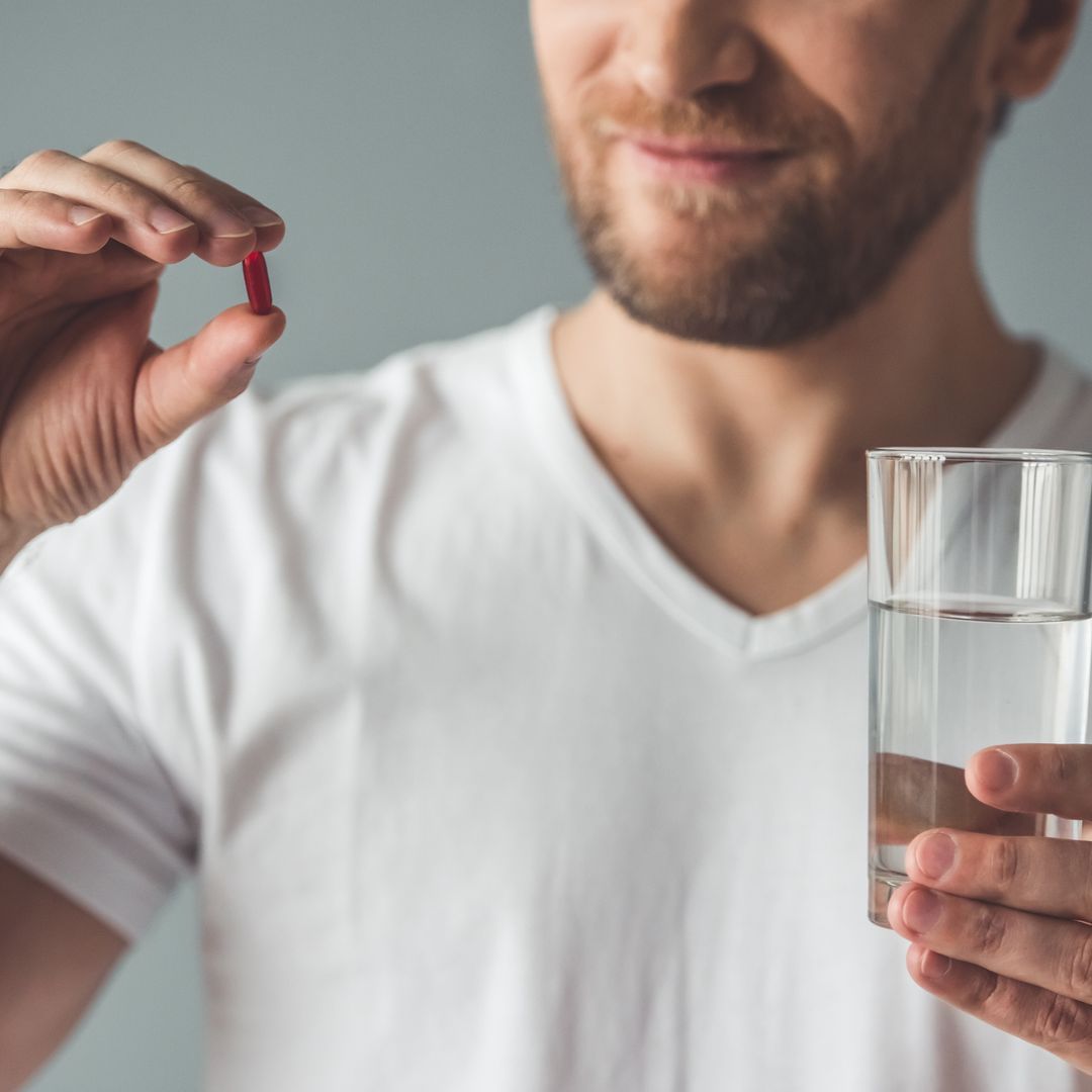 A man holding a supplement capsule and a glass of water in front of a grey background