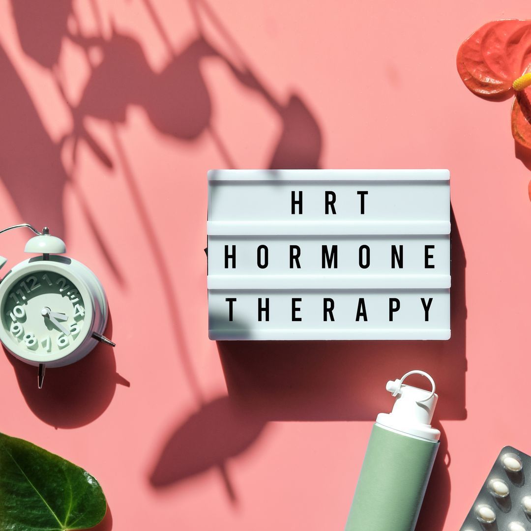 HRT text on a lightbox with an alarm clock, exotic leaves, pills, and oestrogen gel on pink background.