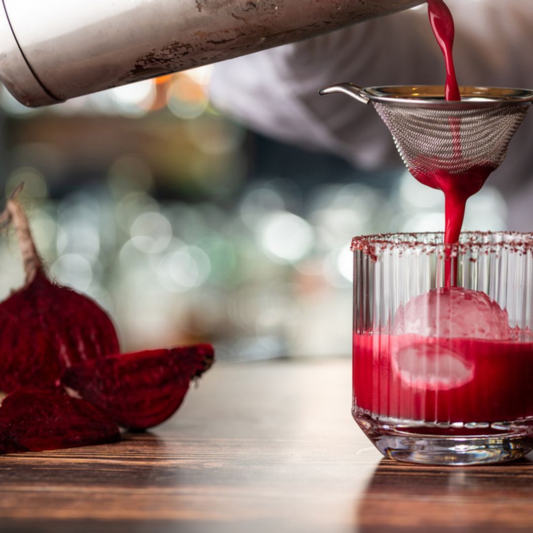 Beetroot juice being poured into a glass with ice through a small metal strainer.