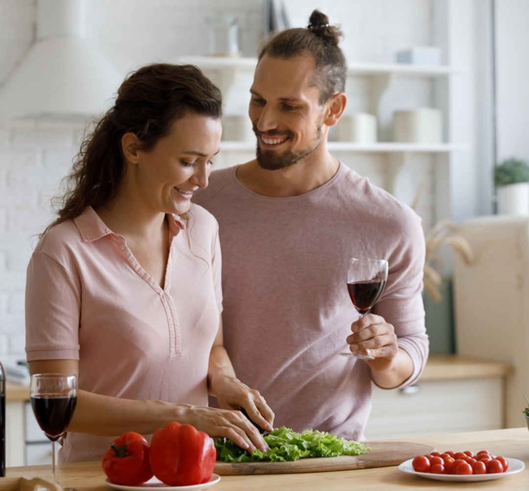 A couple in a kitchen preparing a healthy meal with a glass of red wine.