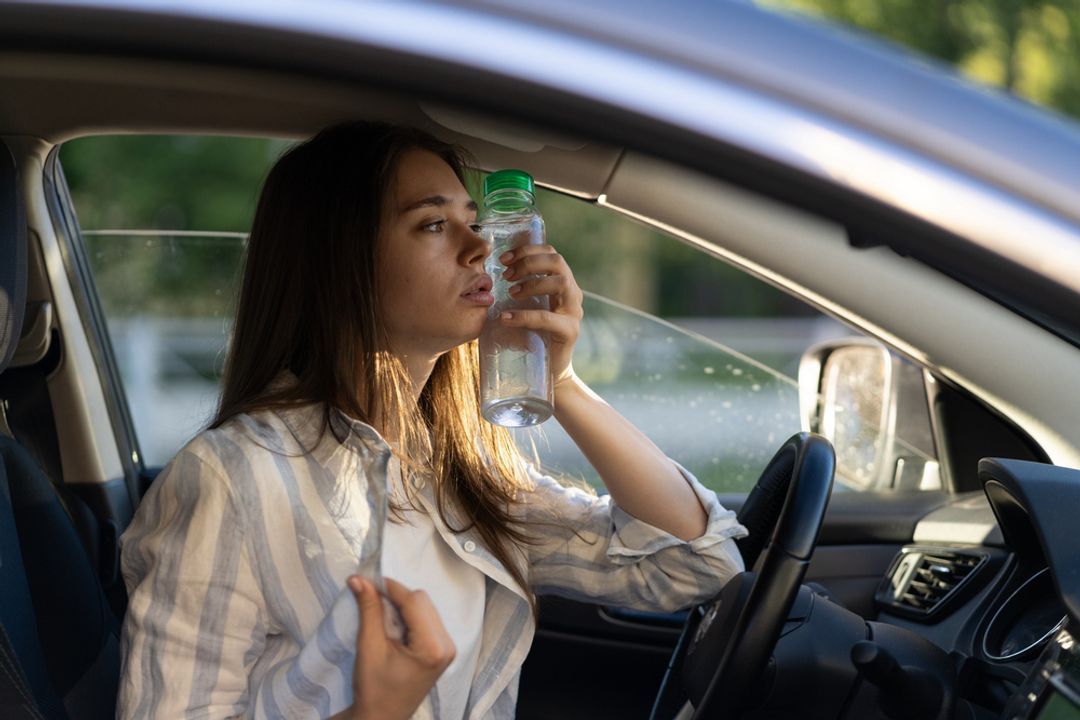 Woman in a car struggling with a hot flash holding a bottle of water to her cheek.  
