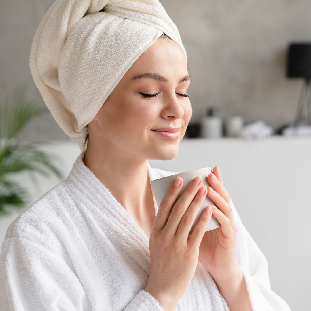 A woman sitting down and enjoying a hot drink in a towel robe with her hair wrapped in a towel