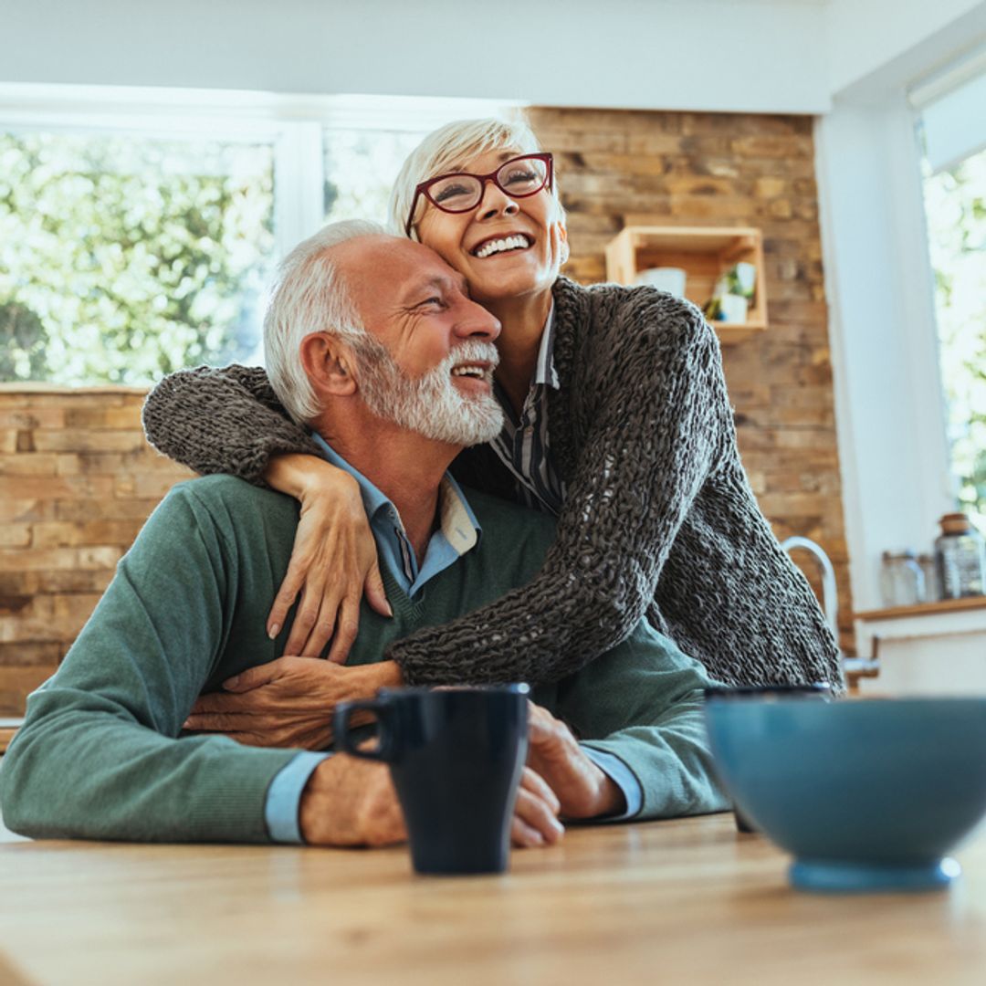 A mature woman and man hugging at the kitchen table smiling.