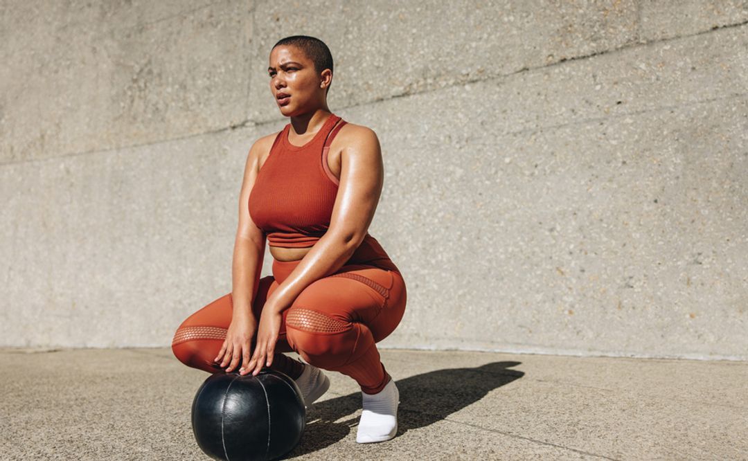  A woman crouching with a medicine ball on the ground outside exercising and looking into the distance.